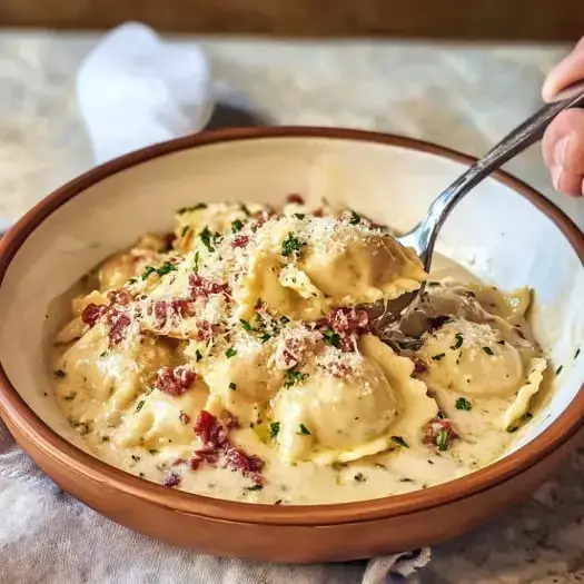 A bowl of velvety ravioli garnished with bacon pieces and parsley, with a fork pulling some pasta up.