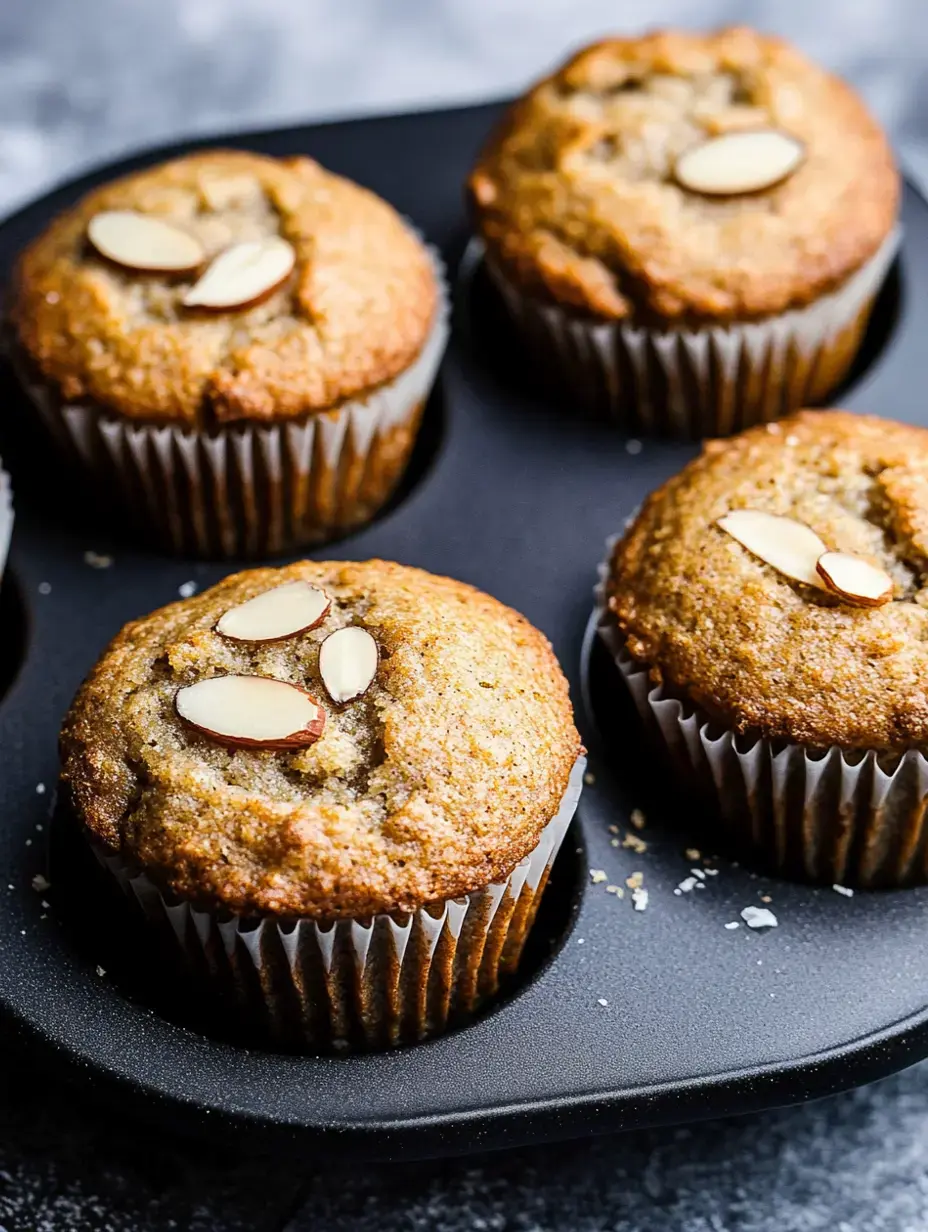 Four fresh almond muffins sit in a black baking tray, sprinkled with sliced almonds on their tops.