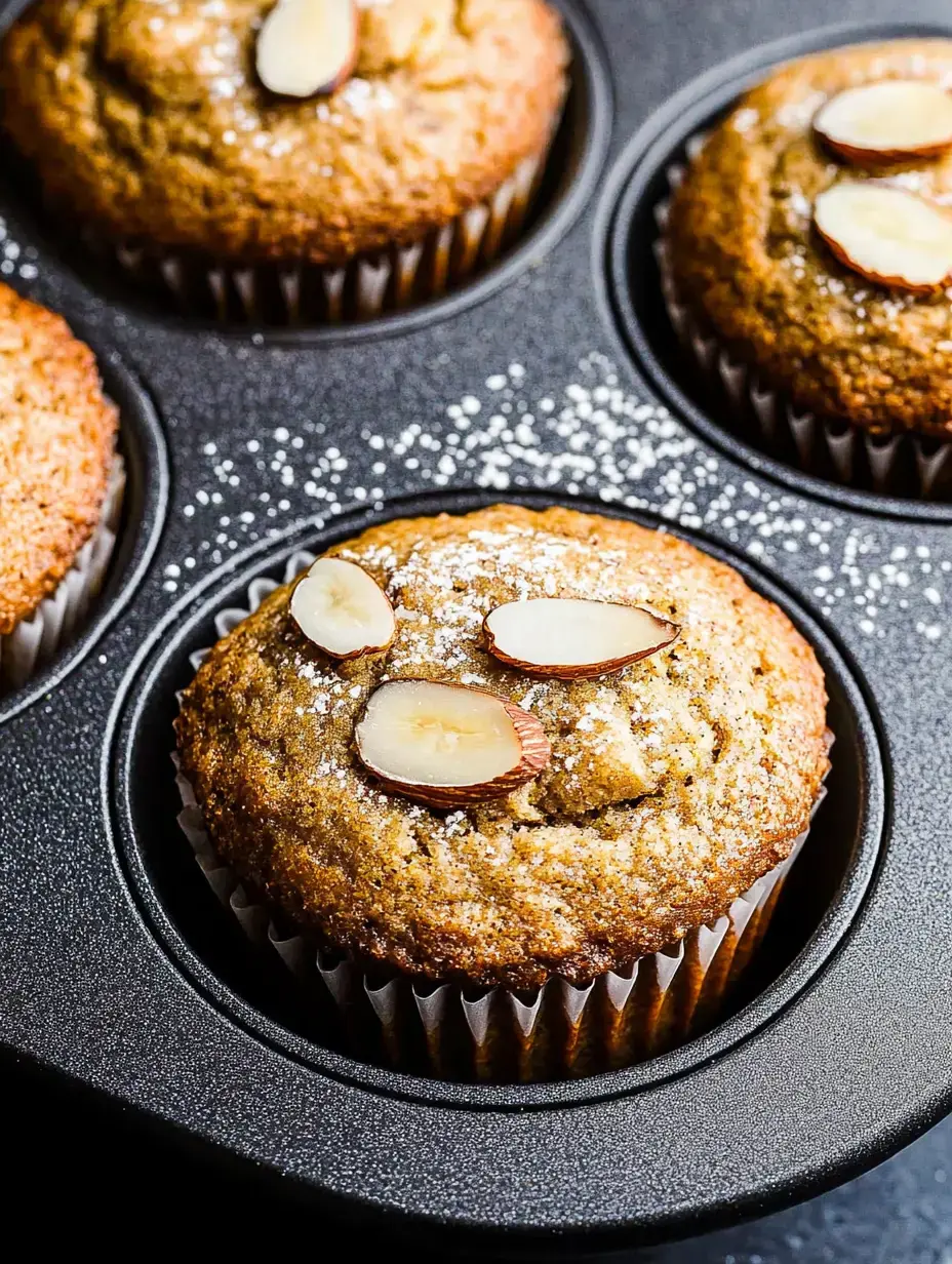 A closeup shot of muffins with almond slices and sugar dusting on top, sitting in a dark-colored baking tin.