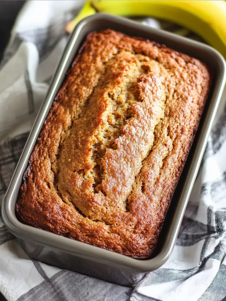A newly baked banana loaf rests in a gray tin, surrounded by ripe bananas and a decorative kitchen cloth.
