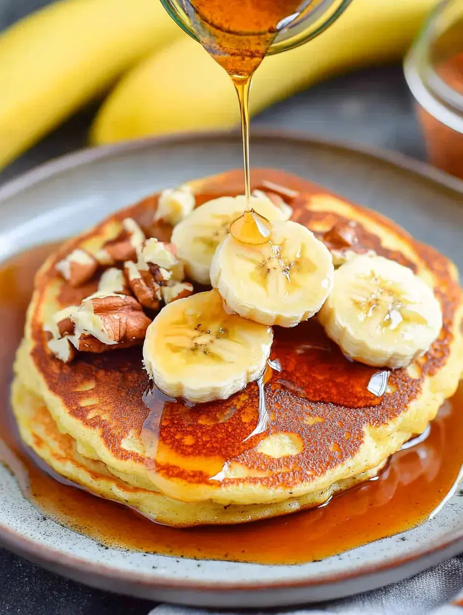 Pancakes stacked together topped with banana pieces, nuts and syrup drizzled over them on a plate.