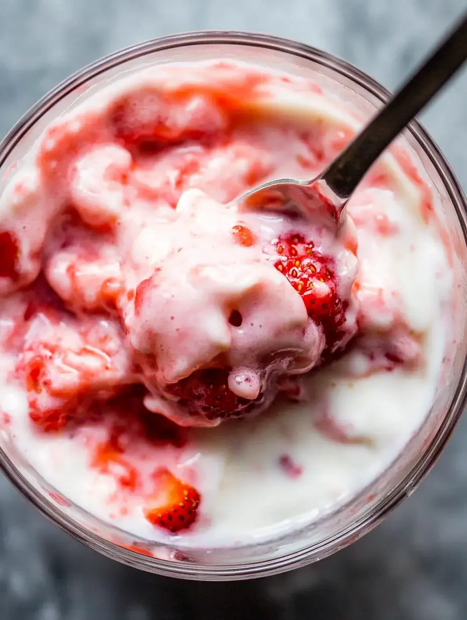 A bowl displays swirled pink yogurt mixed with chunks of fresh strawberries with a metal spoon nestled inside.