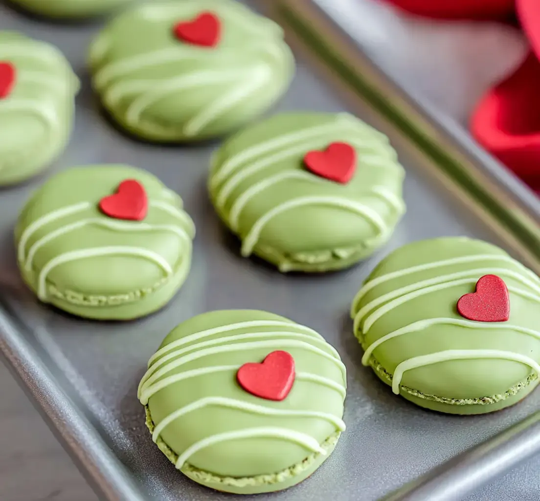 Green sandwich cookies with red heart sprinkles and a drizzle of white chocolate lining a tray.